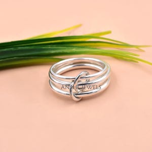 May include: A polished silver ring featuring three interconnected bands that overlap at a central point. The metal surface is smooth and reflective. The text ANTHJEWELS is engraved on the side of the ring band, visible in the center of the design.