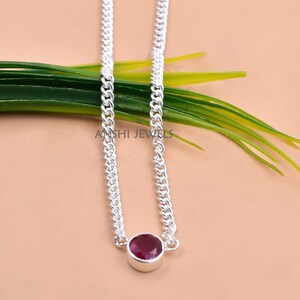 May include: A silver chain necklace featuring a single round, faceted deep red gemstone set in a smooth bezel setting. The necklace is positioned vertically, with the gemstone centred at the bottom of the chain. The text ANSHI JEWELS is visible.