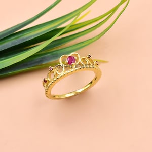 May include: A gold-colored ring in the shape of a crown, featuring a large, round, red gemstone at the center and smaller red gemstones. The ring is on a light pink surface with green leaves in the background. The ring has the text "ANSHI JEWELS".