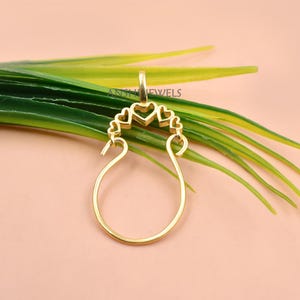 May include: A gold-toned pendant featuring a large circular base with an open center, topped by a decorative arch of five small heart-shaped cutouts. The piece includes a small bail at the top and the text ANSHJEWELS printed in black.