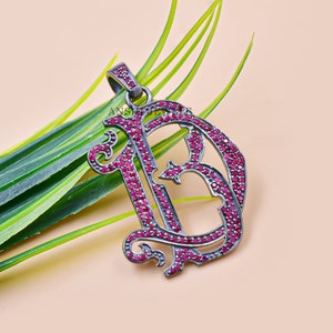 Puede incluir: Un colgante de plata en forma de la letra D, incrustado con piedras preciosas rojas. El colgante está sobre un fondo verde.