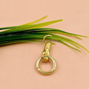 Puede incluir: Un cierre dorado con forma de mano que sostiene un anillo. El cierre está unido a un gancho. El fondo es de color melocotón claro con hojas verdes y amarillas.