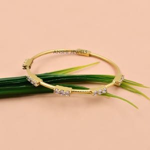 May include: A gold-toned bangle bracelet featuring a textured surface and four rectangular stations, each set with three clear, round-cut stones. The text ANSHI JEWELS is engraved on the metal band. The bangle has a thin, circular design.