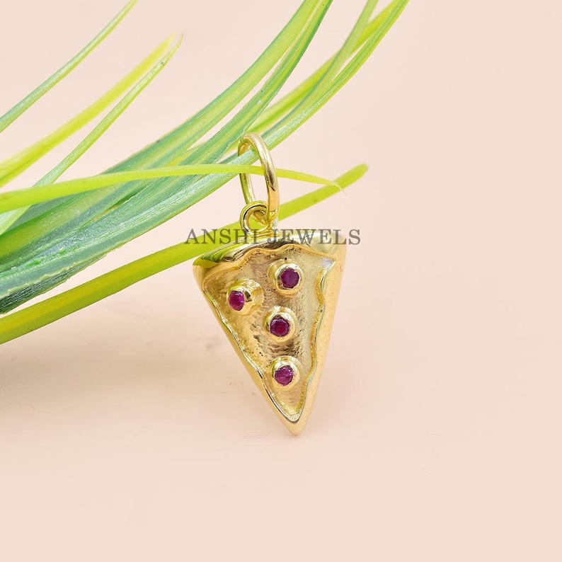 Pu&ograve; includere: Un ciondolo color oro a forma di fetta di pizza, con quattro piccole gemme rotonde e rosse. Il ciondolo ha una superficie strutturata e un anello per una catena. Il testo "ANSHI JEWELS" &egrave; visibile.