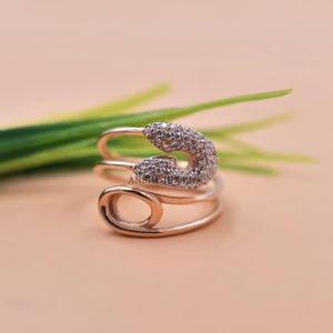 Op de afbeelding: Een ring van roségoud met een veiligheidsspeldontwerp. De speld is bezet met heldere zirkonia-stenen.