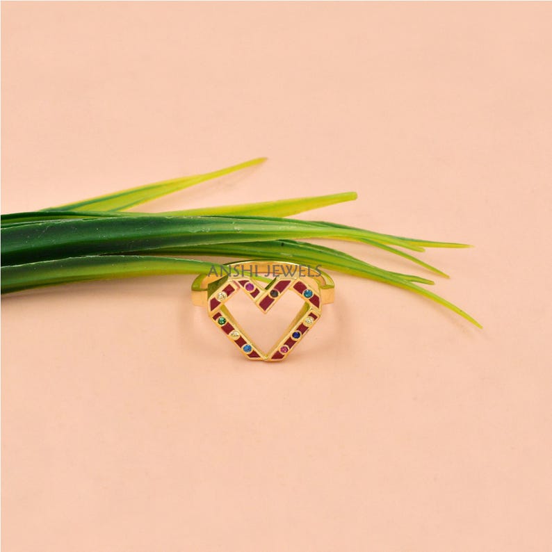May include: A gold ring with a heart-shaped design. The heart is outlined with red and features small, colorful gemstones. The ring is set against a soft pink background with green leaves.