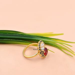 K&ouml;nnte beinhalten: Ein goldener Ring mit einem zentralen ovalen wei&szlig;en Stein und einem runden roten Stein, umgeben von einem Kranz aus kleinen blauen Steinen in einer dekorativen Goldfassung. Der Ring hat eine d&uuml;nne, polierte Goldschiene und liegt vor gr&uuml;nen Bl&auml;ttern.