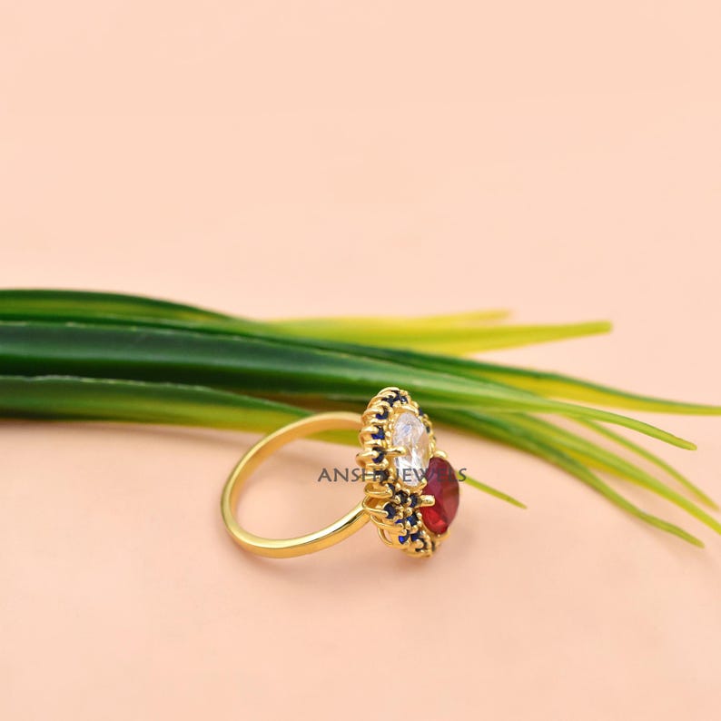 K&ouml;nnte beinhalten: Ein goldener Ring mit einem zentralen ovalen wei&szlig;en Stein und einem runden roten Stein, umgeben von einem Kranz aus kleinen blauen Steinen in einer dekorativen Goldfassung. Der Ring hat eine d&uuml;nne, polierte Goldschiene und liegt vor gr&uuml;nen Bl&auml;ttern.