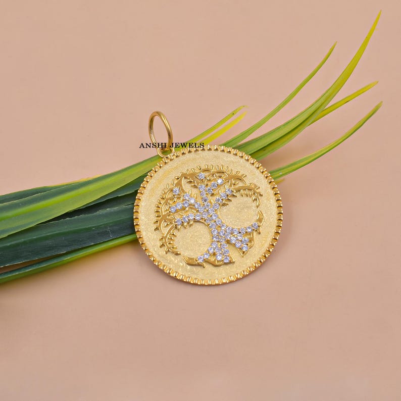 May include: Gold-toned pendant with a tree of life design, embellished with sparkling stones. The pendant has a textured background and a beaded border. The brand name "ANSHI JEWELS" is visible. The pendant is displayed with green leaves against a neutral background.