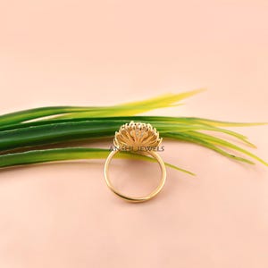 K&ouml;nnte beinhalten: Ein Goldring mit einer kreisf&ouml;rmigen Fassung, einem zentralen klaren Stein und einem Kranz aus kleineren Steinen, der einen Strahlenkranz-Effekt erzeugt. Die Ringschiene ist d&uuml;nn und aus poliertem Gold. Der Text ANSHI JEWELS ist auf dem Ring zu sehen.