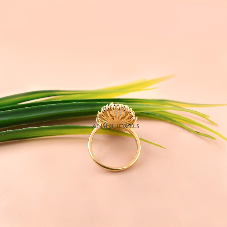 K&ouml;nnte beinhalten: Ein Goldring mit einer kreisf&ouml;rmigen Fassung, einem zentralen klaren Stein und einem Kranz aus kleineren Steinen, der einen Strahlenkranz-Effekt erzeugt. Die Ringschiene ist d&uuml;nn und aus poliertem Gold. Der Text ANSHI JEWELS ist auf dem Ring zu sehen.