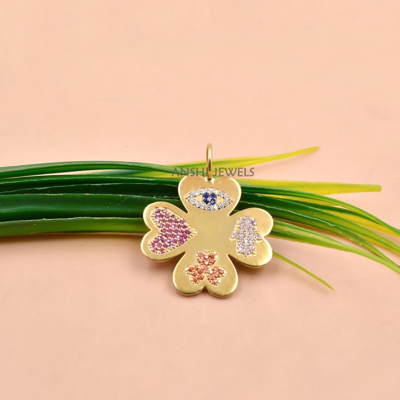 May include: A gold-toned four-leaf clover pendant featuring inlaid gemstones. The design includes a blue and white evil eye, a pink heart, a white hamsa hand, and an orange shamrock. The text ANSHI JEWELS is printed on the metal surface.