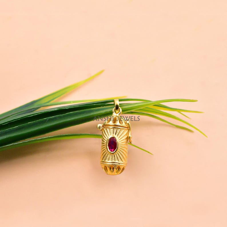 May include: A gold-colored pendant with a cylindrical shape and a red gemstone in the center. The pendant has a sunburst design and is suspended from a small loop. The background is a soft peach color with green leaves.