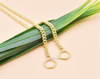 May include: Two gold-coloured chain necklaces with circular pendants are displayed. The chains rest on a long, green leaf against a light peach background. The jewellery is simple and elegant.