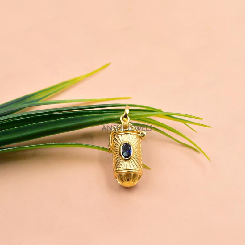 May include: A gold-colored pendant with a cylindrical design and a blue gemstone in the center. The pendant has a sunburst pattern around the gemstone and is suspended from a small loop. The text "ANS Jewels" is visible.