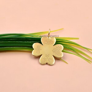 May include: A gold-toned four-leaf clover pendant featuring a smooth centre and textured, dotted patterns on each of the four petals. The pendant includes a small top loop for attachment and displays the text ANSHIJEWELS across the centre.