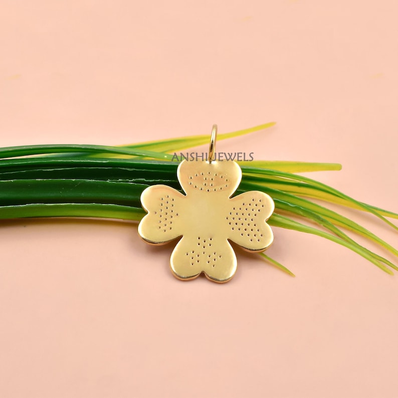 May include: A gold-toned four-leaf clover pendant featuring a smooth centre and textured, dotted patterns on each of the four petals. The pendant includes a small top loop for attachment and displays the text ANSHIJEWELS across the centre.