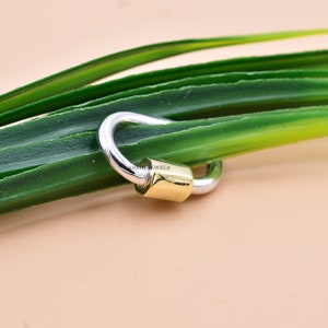 May include: A silver and gold charm with a hook closure. The charm is in the shape of a circle with a small gold cylinder in the centre.