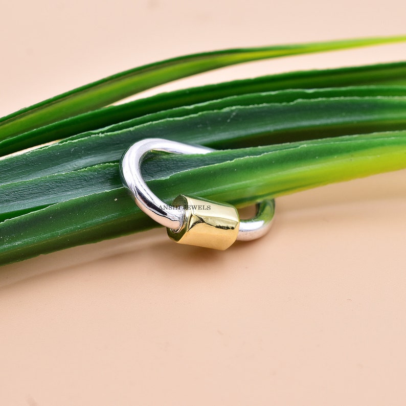 May include: A silver and gold charm with a hook closure. The charm is in the shape of a circle with a small gold cylinder in the centre.