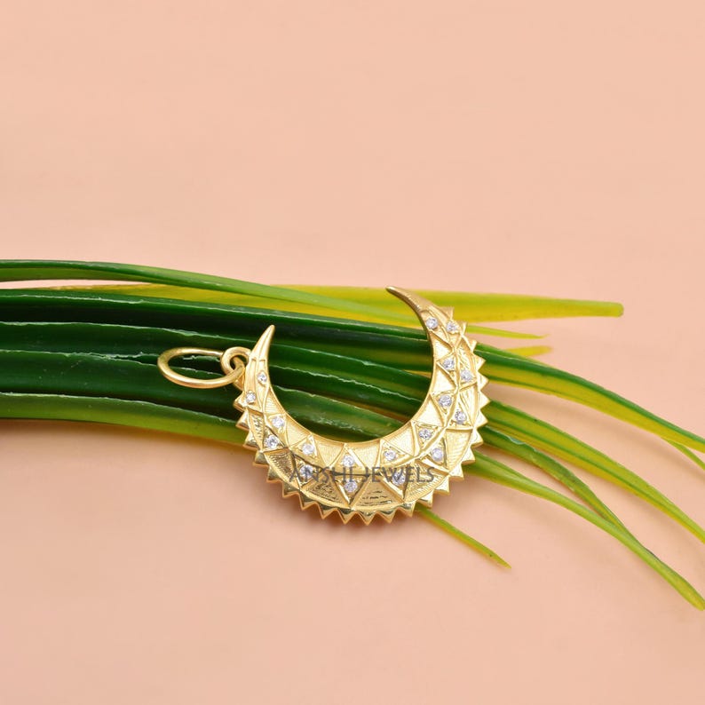 May include: A gold crescent moon pendant featuring a textured geometric pattern with small embedded clear stones and a circular bail at the top. The surface of the pendant displays a repeating triangular design with a polished gold finish.