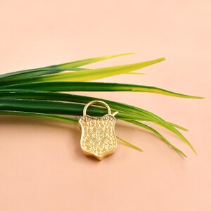 May include: A gold-colored, shield-shaped pendant with an ornate floral design. The pendant has a small loop at the top and is set against a backdrop of green leaves and a peach-colored surface.