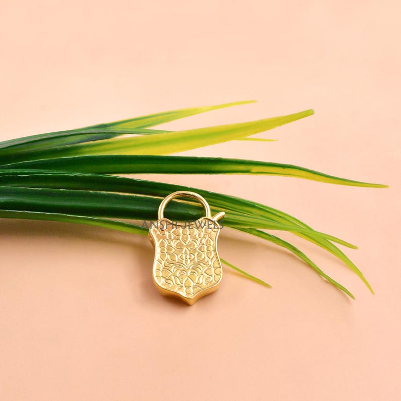 May include: A gold-colored, shield-shaped pendant with an ornate floral design. The pendant has a small loop at the top and is set against a backdrop of green leaves and a peach-colored surface.