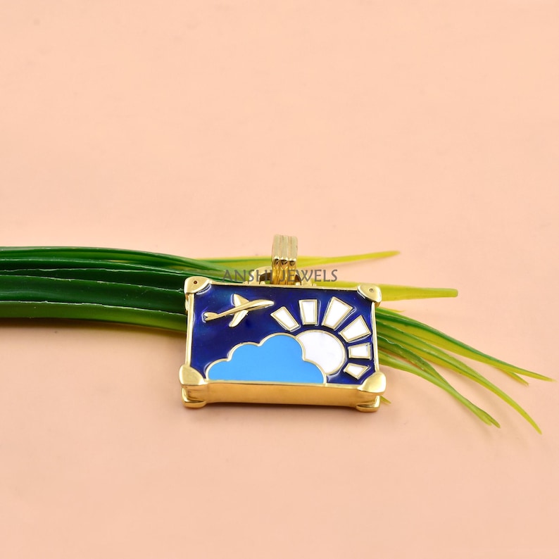 May include: A rectangular gold-tone pendant featuring an enamel design of a blue sky with a white sun, a light blue cloud, and a small airplane. The pendant has a gold bail at the top and the text ANSHJEWELS is visible on the metal surface.