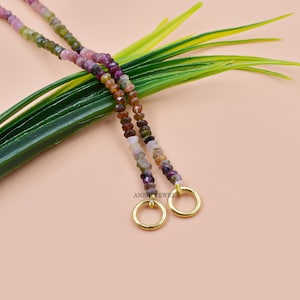 May include: A multi-coloured gemstone beaded necklace with gold-toned end caps. The necklace is made with small, faceted beads in shades of pink, green, brown, and purple. The end caps are circular and have a smooth, polished finish.
