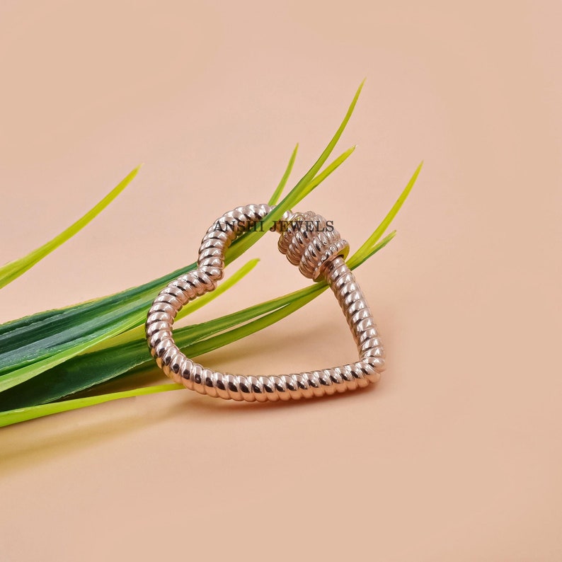 K&ouml;nnte beinhalten: Ein herzf&ouml;rmiger Verschluss in Ros&eacute;gold mit einem strukturierten, seilartigen Design. Der Verschluss ist offen und zeigt die Herzform. Der Hintergrund ist ein weicher Pfirsichton mit gr&uuml;nen Bl&auml;ttern. Der Text "ANSHI JEWELS" ist sichtbar.