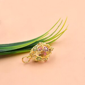 Pode incluir: Um pingente dourado com um design floral, com pedras preciosas verdes e vermelhas. O pingente tem uma argola para uma corrente e est&aacute; sobre um fundo p&ecirc;ssego com folhas verdes.