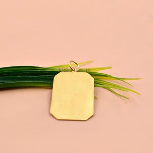 May include: A gold-toned rectangular pendant with clipped corners and a small circular bail at the top. The surface is smooth and solid, featuring the text ANSHI JEWELS printed in black across the centre of the metal plate.