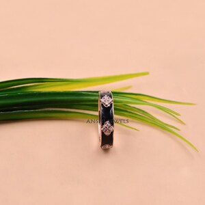 May include: A rose gold band ring featuring a black enamel inlay and a series of four-petaled flower motifs set with small clear stones. The ring is marked with the text ANSHIJEWELS and displays a repeating pattern of these floral accents.