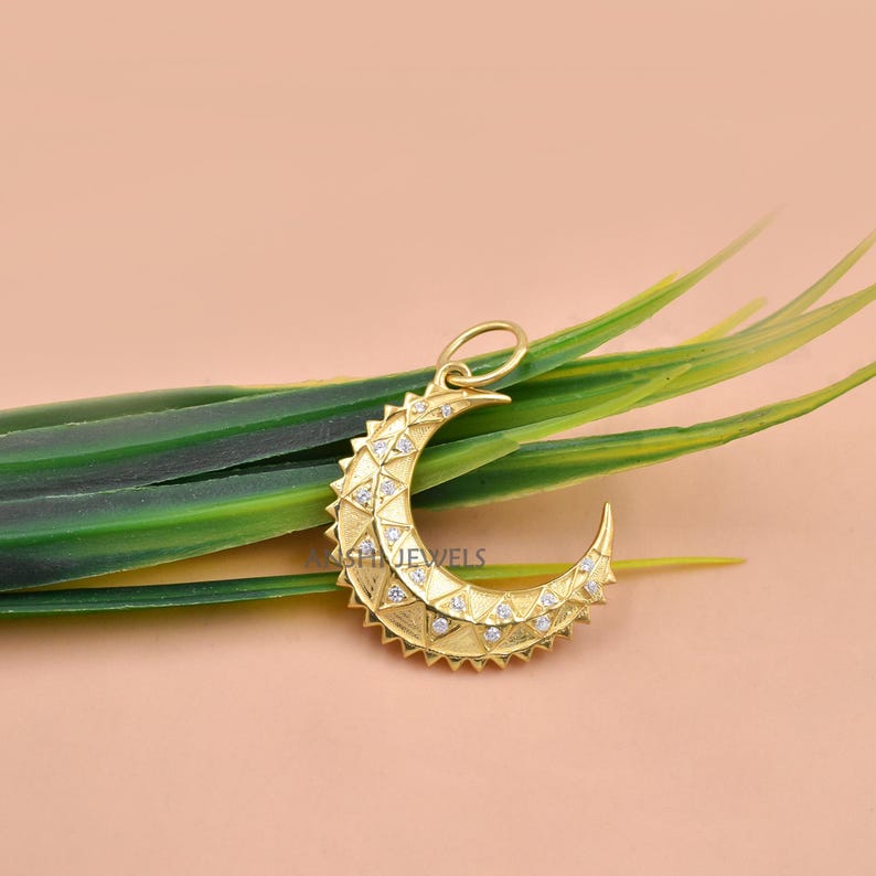 May include: A gold crescent moon pendant featuring a textured geometric pattern across its surface, accented with small embedded clear stones and a circular bail at the top for attachment to a chain or cord, showcasing a detailed celestial design.