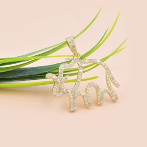 Op de afbeelding: Een gouden olifantenhanger met een diamantbezet ontwerp. De hanger ligt op een groene, bladerige achtergrond.