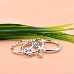 May include: A silver ring featuring an interlocking design with multiple circular bands connected together. A small, clear, square-cut gemstone is suspended from one of the links. The metal has a polished, reflective finish throughout the piece.