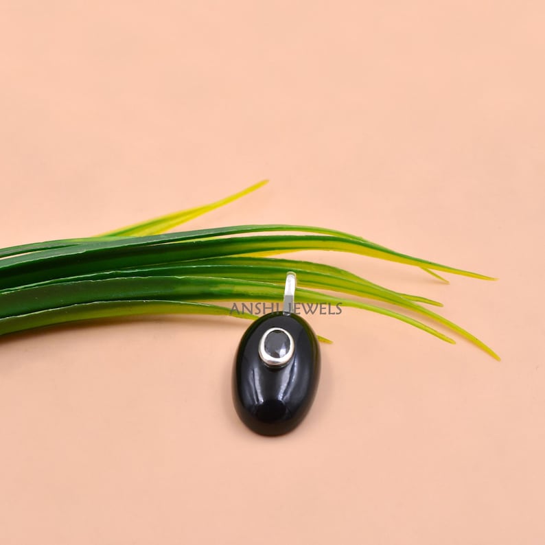 May include: An oval black pendant featuring a small, centered oval gemstone set in a silver-toned metal frame. The pendant has a smooth, polished surface and includes a small silver-toned bail at the top for attachment to a chain or cord.