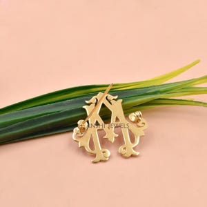 K&ouml;nnte beinhalten: Eine Brosche aus goldfarbenem Metall in Form eines verzierten, stilisierten Buchstabens M mit dekorativen Schn&ouml;rkeln. Das St&uuml;ck hat eine polierte Oberfl&auml;che und einen Ansteckmechanismus auf der R&uuml;ckseite. Der Text ANSHI JEWELS ist sichtbar.