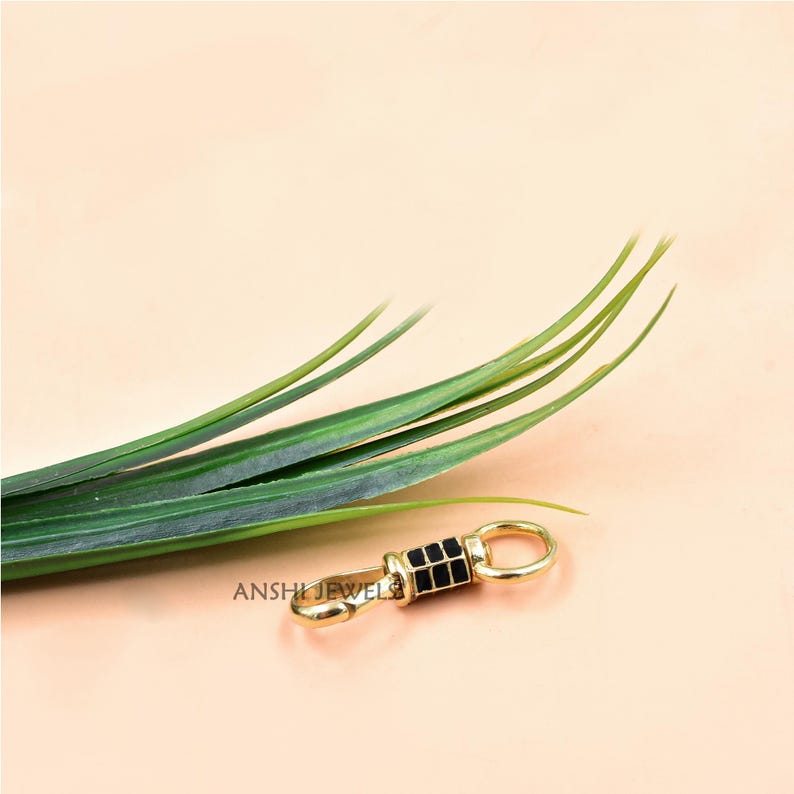 May include: A gold-colored clasp with a black rectangular design is displayed on a light peach background. The clasp has a small ring on one end and a hook on the other. Green leaves are in the background. The text "ANSHI JEWEL" is visible on the clasp.
