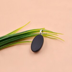 May include: A smooth, oval-shaped black pendant featuring a silver-toned metal bail at the top. The pendant is positioned against a backdrop of green grass-like blades, with the text ANSHI JEWELS faintly visible on the surface of the black stone.