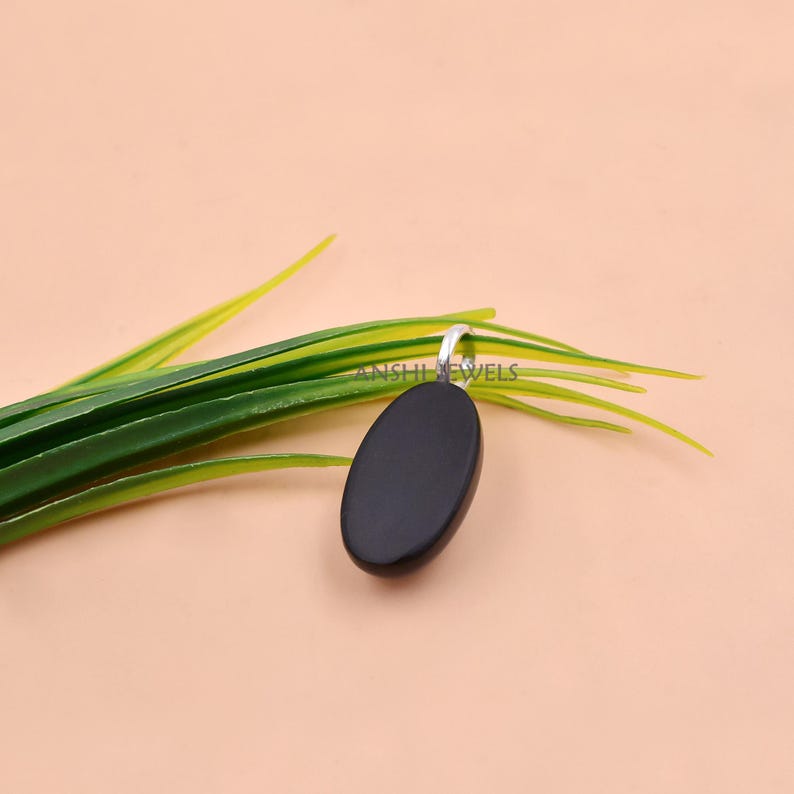 May include: A smooth, oval-shaped black pendant featuring a silver-toned metal bail at the top. The pendant is positioned against a backdrop of green grass-like blades, with the text ANSHI JEWELS faintly visible on the surface of the black stone.