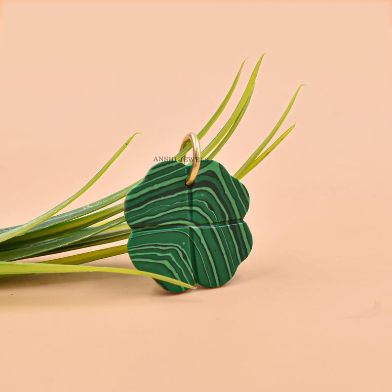 May include: A four-leaf clover pendant with a gold-colored loop. The pendant is green with black veining, resembling malachite. The background is a light peach color with green grass.