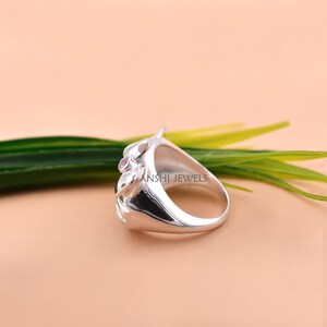 May include: A polished silver ring featuring a small, round, light pink gemstone set into a smooth, curved metal band. The side of the ring is engraved with the text ANSHI JEWELS, showcasing a reflective surface and a rounded, organic design.
