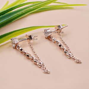 Puede incluir: Pendientes de oro rosa con dise&ntilde;o de cadena. Cada pendiente presenta una gema transparente de corte cuadrado y una cadena con eslabones decorativos. El texto "ANSHI JEWELS" es visible. Sobre una superficie rosa claro.