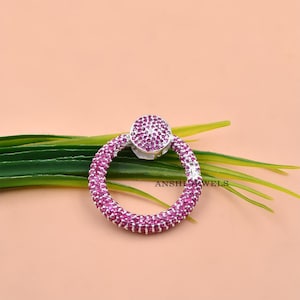 Puede incluir: Un colgante circular con una base de metal plateado totalmente incrustada con pequeñas piedras de color rosa vibrante. La pieza incluye una argolla de conexión superior también cubierta de piedras rosas. El texto ANSHI JEWELS es visible.