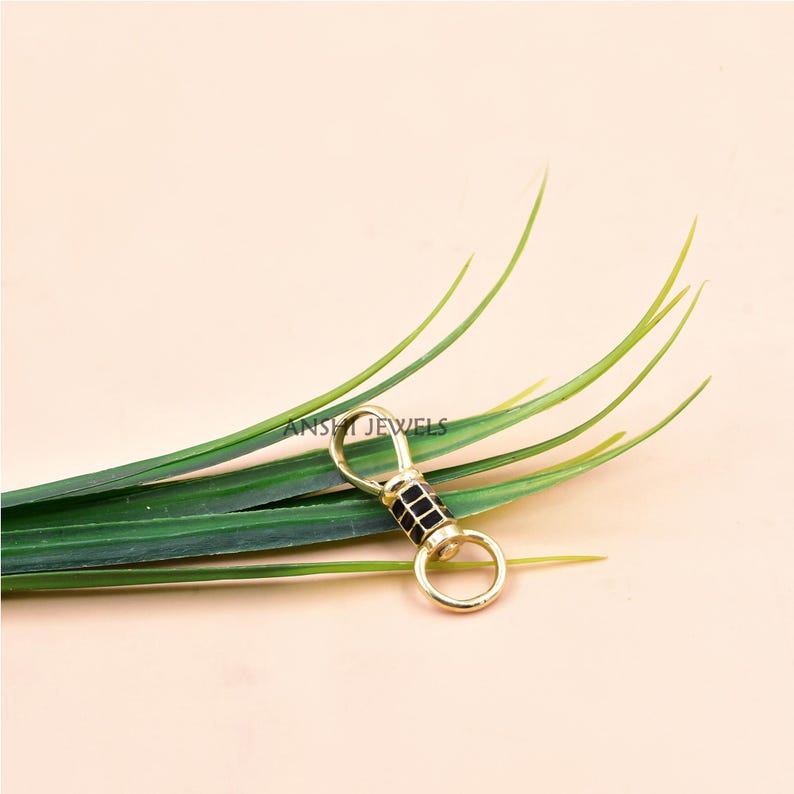May include: A gold-colored keychain clasp with a black, square-patterned center. The clasp has a circular ring at each end and is set against a backdrop of green leaves and a light pink surface. The text "ANSKI JEWELS" is visible.