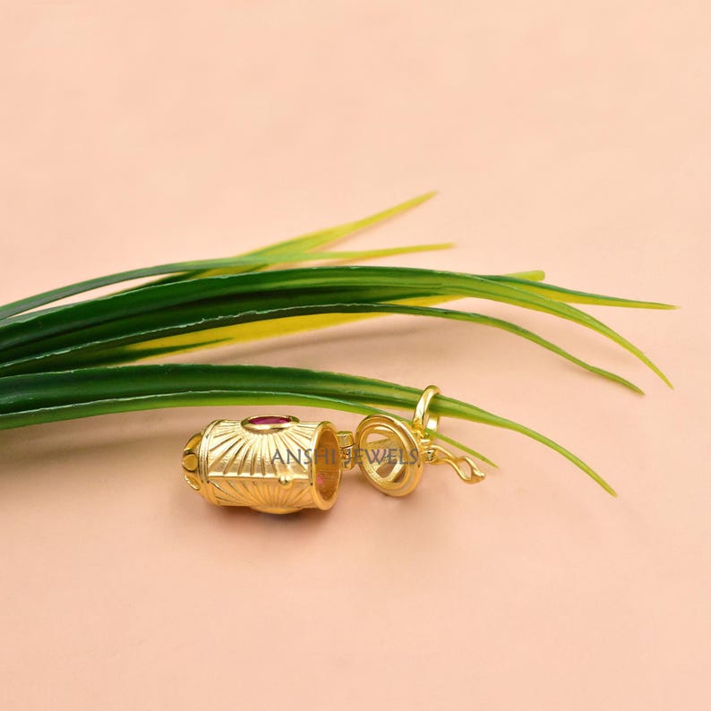 May include: A gold-colored pendant with a cylindrical design, featuring a red gemstone and intricate detailing. The pendant is open, revealing the interior. The text "ANSHI JEWELS" is visible on the pendant. The pendant is set against a light pink background with green leaves.