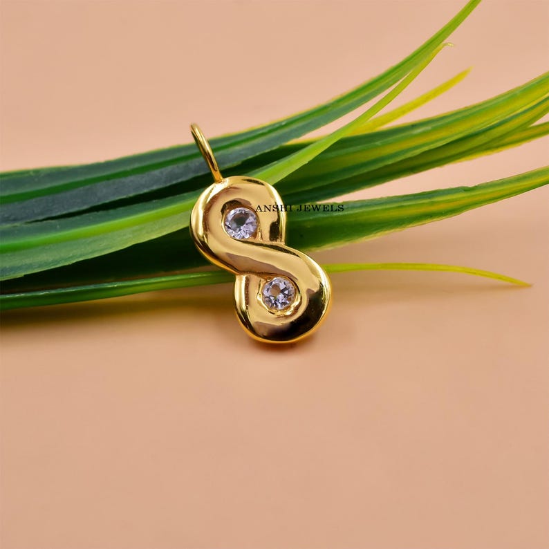 May include: A gold-colored pendant in an infinity symbol shape, featuring two clear gemstones. The pendant is displayed against a backdrop of green leaves and a peach-colored surface. The text "ANSHI JEWELS" is visible.