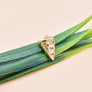 Pu&ograve; includere: Un ciondolo a forma di fetta di pizza color oro con tre pietre preziose rosse. Il ciondolo ha un anello nella parte superiore per attaccarlo a una collana o un braccialetto. Il ciondolo &egrave; posto su uno sfondo di foglie verdi.