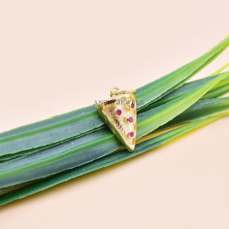 Pu&ograve; includere: Un ciondolo a forma di fetta di pizza color oro con tre pietre preziose rosse. Il ciondolo ha un anello nella parte superiore per attaccarlo a una collana o un braccialetto. Il ciondolo &egrave; posto su uno sfondo di foglie verdi.