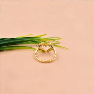 May include: A gold-colored ring in the shape of a heart, with the brand name "ANSHI JEWELS" visible. The ring is set against a soft pink background, with green leaves in the upper left corner.